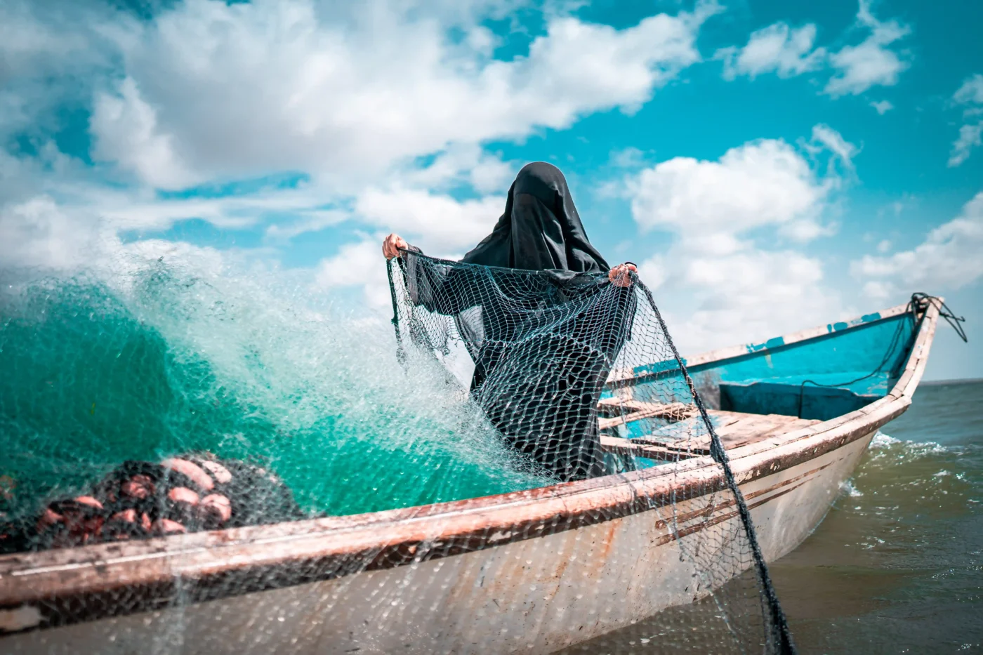A woman dressed in a niqāb casts her net into the water from a fishing boat.