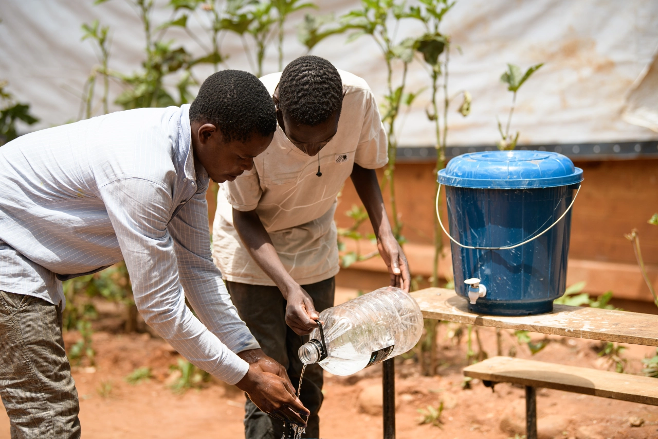 Two men at a hand washing station. One of the men pours the water from a large water bottle over the other man's hands.